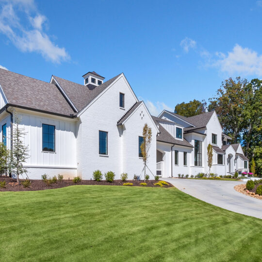 A row of modern white houses with gabled roofs on a sunny day. The houses are aligned along a curving driveway, with a well-manicured green lawn and young shrubs in the foreground. The sky is blue with scattered clouds.