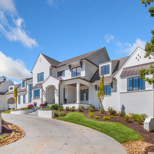 A large, modern white house with multiple gables sits atop a sloped driveway. The home features large windows, manicured landscaping, and a bright, sunny sky overhead.