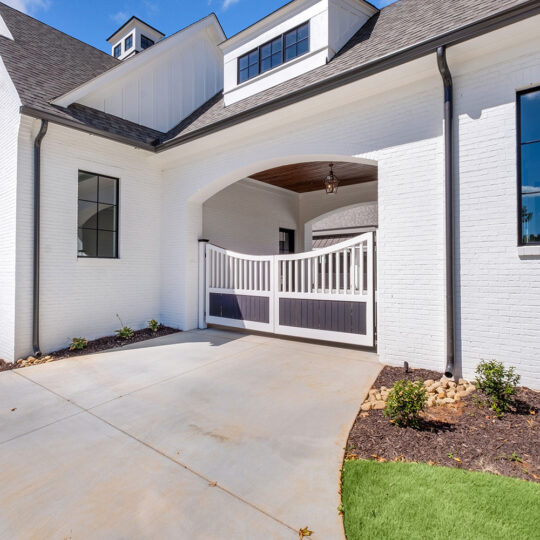 Driveway leading to a white brick house with a dark roof. The house features large black-framed windows and a double gate. The surrounding landscaping includes small shrubs and freshly laid sod. The sky is clear with a few clouds.