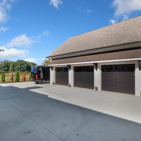 A large, modern garage with three dark brown doors is shown. A black van is parked nearby. A basketball hoop is visible near a fence, with greenery in the background under a blue sky with clouds.