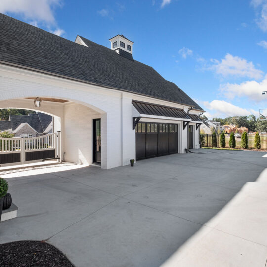 Modern residential driveway with a white garage and black doors on the left side. The building features a dark roof and cupola. A basketball hoop is visible in the background, along with trees and a wooden fence under a clear blue sky.