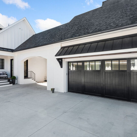 Modern, white two-story house with a large black garage door and spacious driveway. The entry features a small porch with seating and potted plants. The roof is dark gray, and the sky is clear with some clouds.