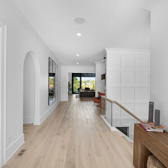 A modern hallway with light wood flooring, white walls, and recessed lighting. Theres a round mirror on the right and a narrow wooden console table below. The end of the hallway leads to a brightly lit room with large windows.