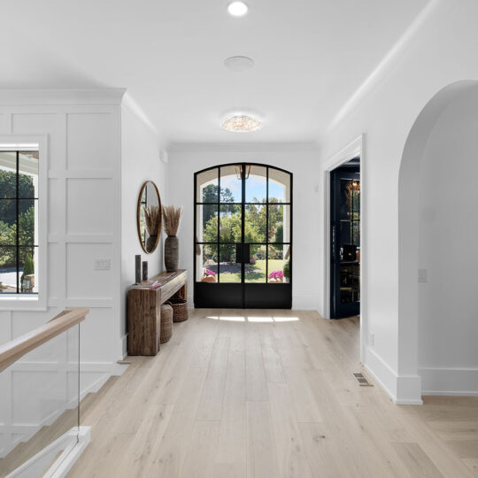 Bright, modern hallway with light wood flooring, a large arched window and door, white walls, and a decorative console table with a round mirror. Natural light streams in, highlighting the contemporary and clean design.