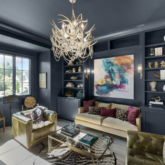 Stylish living room with dark blue walls and ceiling. Features include a modern chandelier, abstract art, shelves with decor, and gold velvet furniture. Large window provides natural light, showcasing a cozy, sophisticated space.