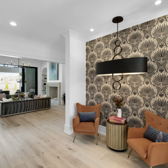 Modern living room with a stylish wallpapered accent wall, featuring two brown chairs and a round table with flowers. A large, dark chandelier hangs above. In the background, a spacious sitting area with a sectional sofa and large windows is visible.