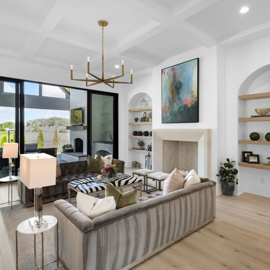 Modern living room featuring a plush gray sofa, large glass doors opening to a patio, and light wood flooring. A fireplace is flanked by built-in shelves with decor. A large abstract painting and chic chandelier complete the space.