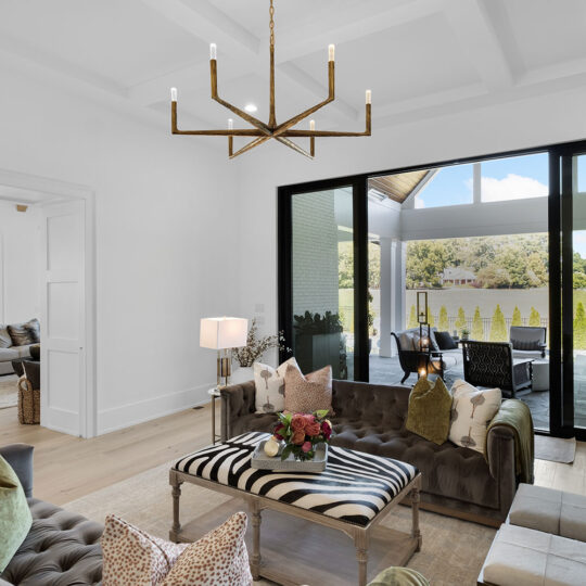 A modern living room with two gray tufted sofas and a zebra print coffee table. Large sliding glass doors open to a patio with outdoor chairs and tables. A geometric chandelier hangs from the ceiling. Neutral decor accents the space.