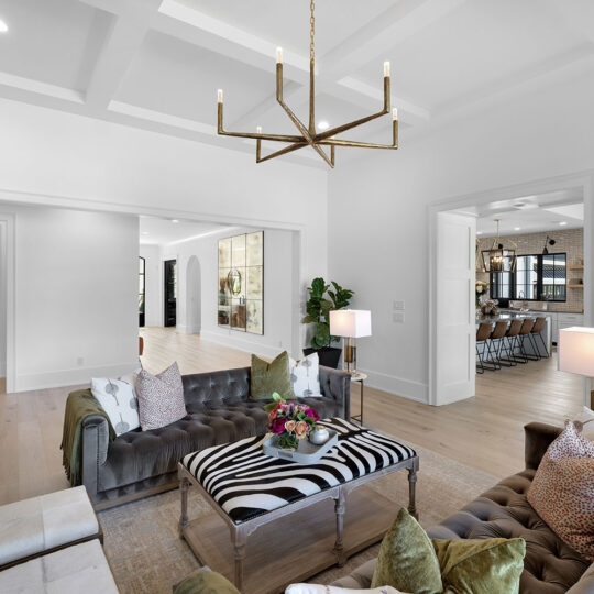 Modern living room with a light wood floor, olive velvet sofas, and a zebra-striped coffee table. A geometric chandelier hangs from the coffered ceiling. Open view to a dining area and a kitchen. Plush pillows and floral centerpiece adorn the space.