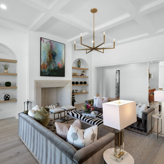 A modern living room with gray couches, a striped ottoman, and decorative pillows. A geometric chandelier hangs from the coffered ceiling. Shelves flank a fireplace with abstract art above. Large windows in an adjoining room let in natural light.