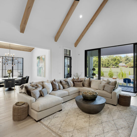 Spacious living room with vaulted ceiling and wooden beams, featuring a large beige sectional sofa and round coffee table on a textured rug. Glass doors open to a view of a lush garden. Adjacent dining area with a modern chandelier and window views.