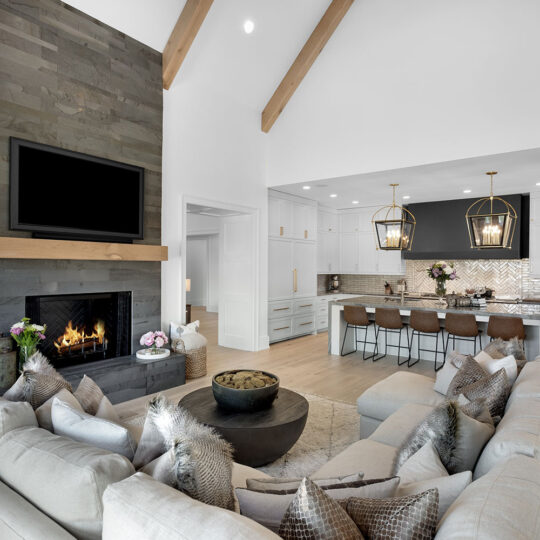 Modern open-concept living room and kitchen with high wooden beams. Features a large sectional sofa, wall-mounted TV, fireplace, and island with bar stools. Kitchen has pendant lights, white cabinets, and a patterned tile backsplash.