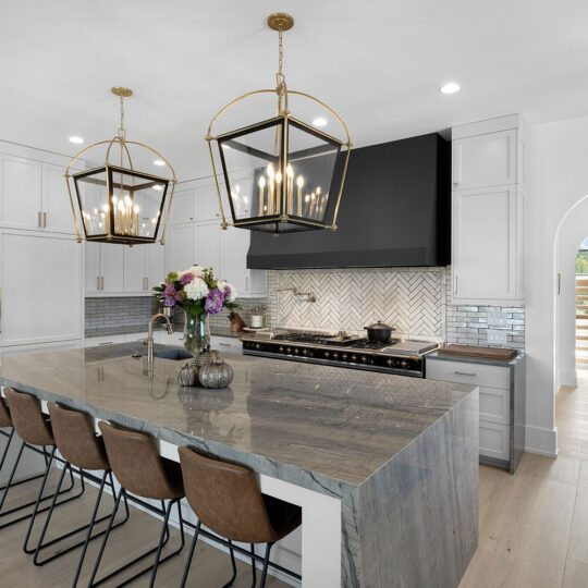 Modern kitchen with a large marble island, five brown barstools, gold-framed pendant lights, and white cabinetry. A stovetop with a dark hood is set against a herringbone-patterned backsplash. An arched doorway leads to a bright hallway.