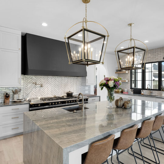 Modern kitchen with a large island featuring a marble countertop and brown leather stools. Pendant lights hang above. White cabinets, a dark stove hood, brick backsplash, and a vase with flowers complete the elegant look.