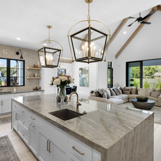 Spacious modern kitchen and living area with large island, pendant lights, exposed wooden beams, and sliding doors opening to a lush garden view. White cabinetry, a cozy seating area, and bright natural light enhance the elegant design.