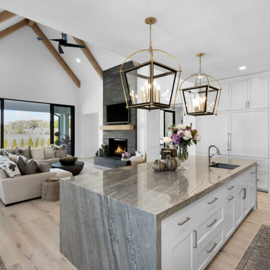 Spacious modern kitchen and living area with a large marble island, brass light fixtures, and a cozy seating area by a fireplace. Vaulted ceiling with wooden beams and large sliding doors open to a scenic outdoor view. Neutral tones with floral decor.