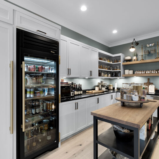 A modern kitchen with white cabinets, a glass-door refrigerator, and an island with wooden countertops. Shelves with jars and bottles line the walls. The space is lit by ceiling and wall lights, creating a cozy atmosphere.