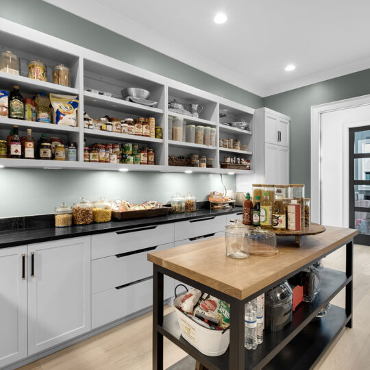 A modern pantry with white shelves filled with jars, cans, and bottles. A wooden-topped island in the center holds various condiments. The room has green walls, light wood floors, and a doorway leading to another room.