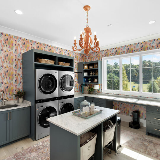 A bright laundry room with teal cabinets and a colorful patterned wallpaper. Theres a stacked washer and dryer, a large window with a scenic view, and an island with storage. A chandelier hangs from the ceiling.