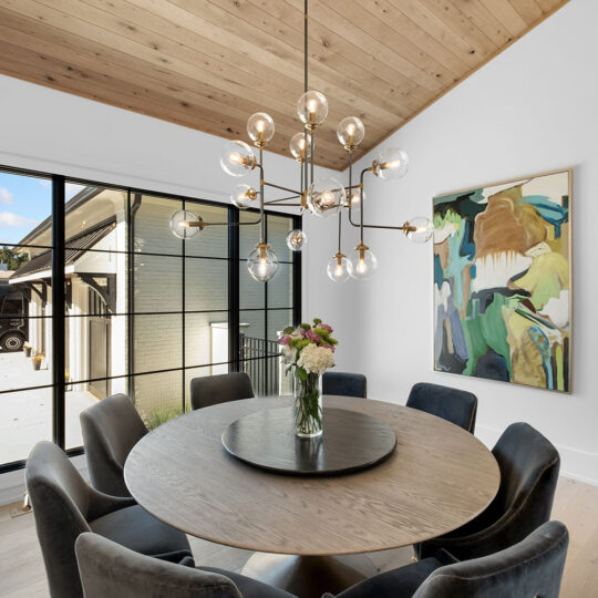 A modern dining room with a round wooden table and dark velvet chairs. A large chandelier with spherical bulbs hangs above. A colorful abstract painting is on the wall, and a large window shows an outdoor view with a patio and parked vehicle.