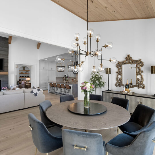 A modern dining room with a round table surrounded by six velvet chairs. A chandelier hangs above. The living room is visible with a sofa and fireplace. A kitchen with bar stools is in the background. The floor is wooden, and a plant is in the corner.