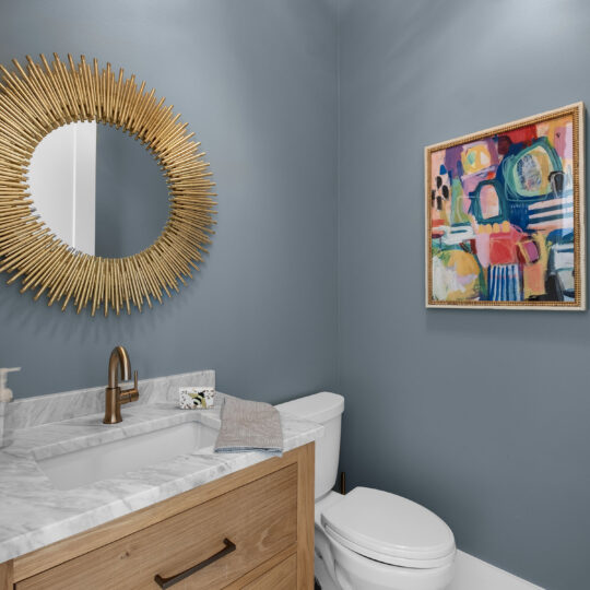 A modern bathroom with blue walls features a round, gold sunburst mirror above a marble sink. Wooden cabinetry and a white toilet are present. A colorful abstract painting adorns the right wall. Light wood flooring completes the decor.