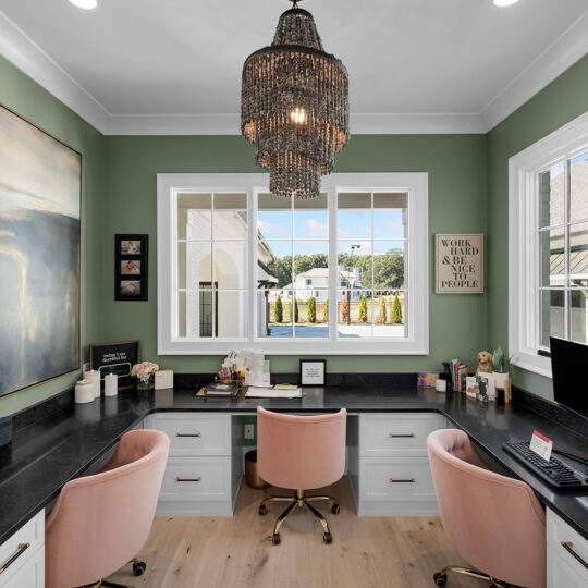 A home office with two light pink chairs at a black L-shaped desk. The walls are green and adorned with a large painting. A window with a view outside is centered between the desks, and a chandelier hangs from the ceiling.