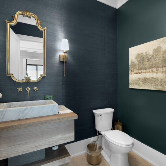 A modern bathroom with dark green walls, a marble sink with a wooden base, and a white toilet. A gold-framed mirror and wall sconce hang above the sink. A painting depicting trees is on the wall. The floor is light-toned wood.