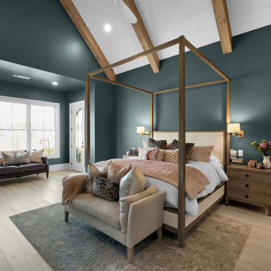 Spacious bedroom with a peaked ceiling and exposed wooden beams. Features a wooden canopy bed, a beige loveseat, a large window with a view, and green walls. Deco pillows and wooden nightstand add cozy accents. Light hardwood floor completes the scene.