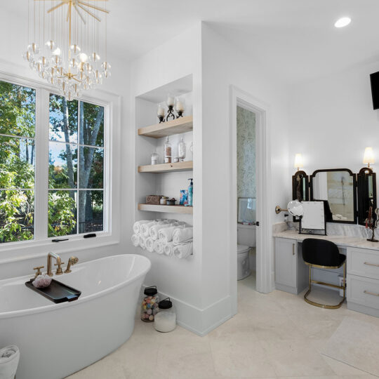 Luxurious bathroom with a freestanding bathtub under a large window, elegant chandelier, and rolled towels on shelves. A vanity with a mirror and chair is on the right, near a wall-mounted TV. Neutral tones and ample natural light enhance the space.