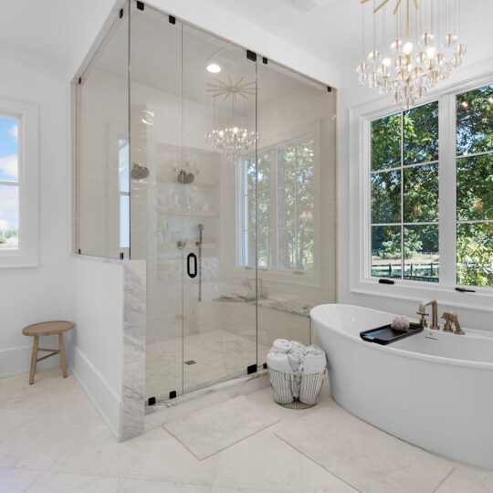 A luxurious bathroom with a freestanding bathtub next to large windows showing a scenic view. A glass-enclosed shower stands nearby, with a stool and rolled towels. A chandelier hangs above, and the room is bright with natural light.