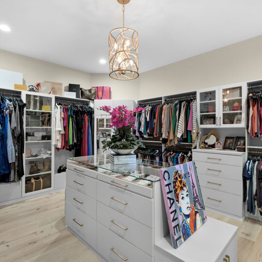 Spacious walk-in closet with organized clothing racks, a central island featuring a glass top and drawers, luxury brand boxes on shelves, and a decorative light fixture. A colorful shopping bag and orchid plant adorn the island.