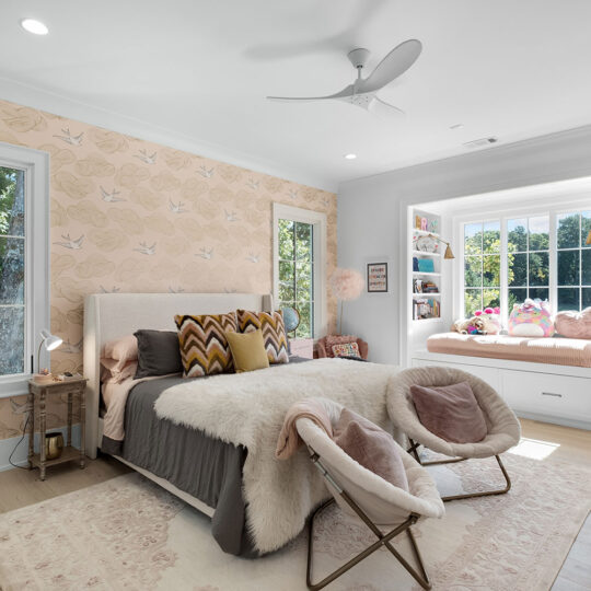 A cozy bedroom with a window seat featuring plush toys, a large bed with pillows and throws, two stylish chairs, and patterned wallpaper. Light streams in through large windows, illuminating the rooms soft, elegant decor.