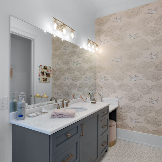 Bathroom with dual sinks, dark gray cabinetry, and a large mirror. The wall has decorative bird-patterned wallpaper. Two sets of lights hang above the mirror. A round, textured rug lies on the hexagonal-tiled floor.