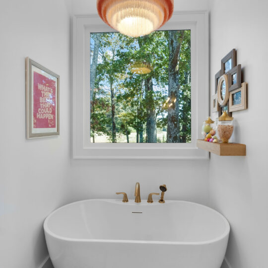 A modern bathroom features a white freestanding bathtub under a large window showing trees. A gold faucet and a round pendant light hang above. A framed quote and small decorations are on the wall, with towels in a basket on the floor.