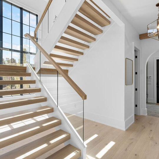 A modern, bright interior featuring a wooden staircase with glass railing. Large windows light the space, and a geometric pendant light hangs from the ceiling. A hallway leads to a doorway with a view outside. Light wood floors complement the decor.