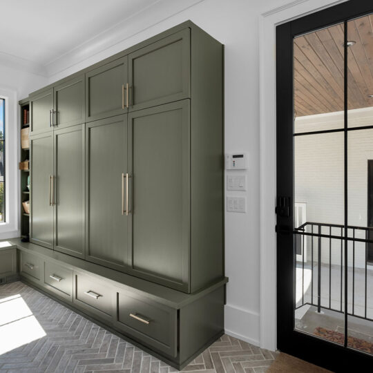 A modern hallway features a large olive green closet with gold handles. Sunlight streams through a window onto the herringbone tile floor. A glass door with a black frame leads outside to a porch with wooden ceiling panels.