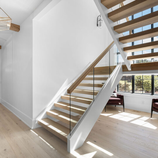 Bright modern hallway with light wood flooring and a staircase featuring glass and wood railings. Two armchairs and a plant are near large windows, allowing natural light. Geometric pendant lights hang from the ceiling.