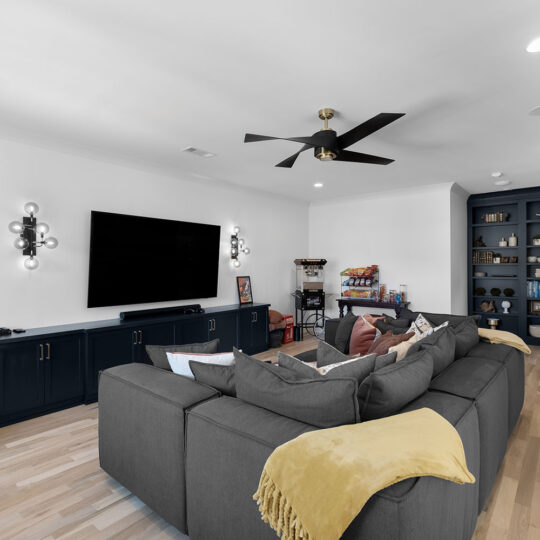 Modern living room with a large gray sectional sofa, yellow throw blankets, a flat-screen TV, and black entertainment unit. Built-in shelves filled with decor and a ceiling fan add to the cozy atmosphere. Light wood flooring and soft lighting complete the space.