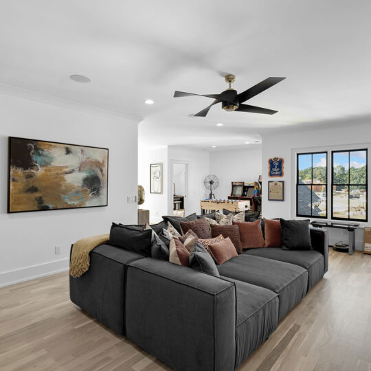 Contemporary living room with a large gray sectional sofa accentuated by an array of cushions. A ceiling fan hangs above. Modern artwork decorates the walls. Sunlight streams through large windows revealing a suburban view. Boxes are stacked in the corner.