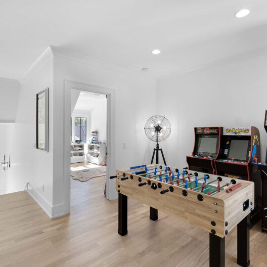 Modern game room with foosball table and three arcade machines against a white wall. A large, round studio light stands nearby. The room has light wood flooring and a doorway leading to another room with a sofa and large window.