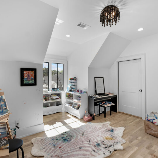 A bright art studio with a sloped ceiling features an easel, art supplies, and paintings. A keyboard sits near a window, and a plush bean bag and rug are on the wooden floor. A unique chandelier hangs from the ceiling.