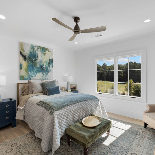 A bright bedroom with a large bed featuring layered pillows and a textured blanket. A green ottoman is at the foot. A blue dresser with lamps and photos is on the side. An armchair is in the corner. Large windows offer a view of greenery outside.