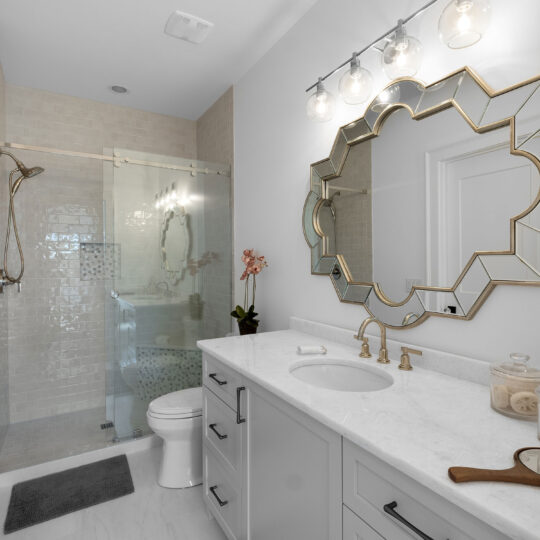 Elegant bathroom with a white vanity, gold faucet, and a decorative mirror. Glass shower enclosure with rainfall showerhead. Soft lighting from globe fixtures above the mirror. Gray floor mats and a small orchid plant accent the clean space.