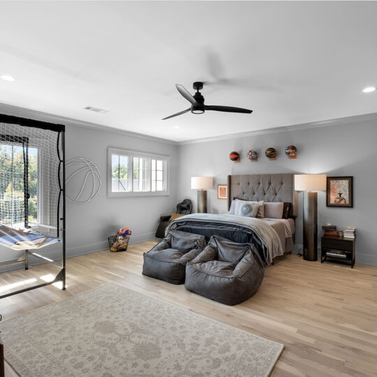 A modern bedroom with a large bed, gray headboard, and blue bedding. Two bean bags rest at the foot of the bed. A desk with a table tennis net is by the window. Masks decorate the wall above the bed. Light wood floors and neutral walls complete the look.