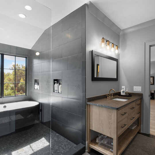 Modern bathroom with dark tiled walls and large window, featuring a freestanding bathtub and walk-in shower. Wooden vanity with a black countertop and a rectangular mirror above. Open door leads to a bright room with a dog bed and gym equipment.