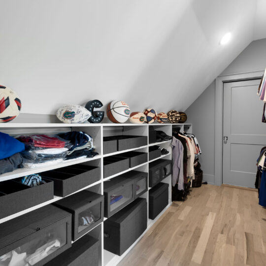 A well-organized closet with wooden floors, featuring shelves with folded clothes, black storage boxes, and neatly hung clothes. Sports balls, including soccer and basketballs, are displayed on the top shelf. A door is visible at the end.