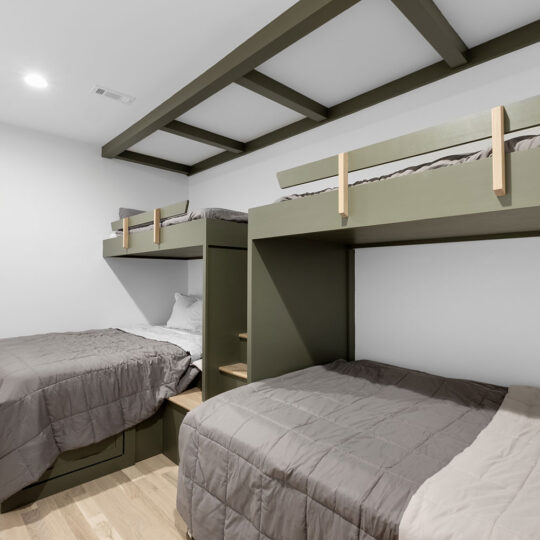 A modern bedroom with four olive green built-in bunk beds. Each bed is neatly made with grey bedding. The room features light wood floors and a white wall, with an exposed beam ceiling in the same shade as the beds.