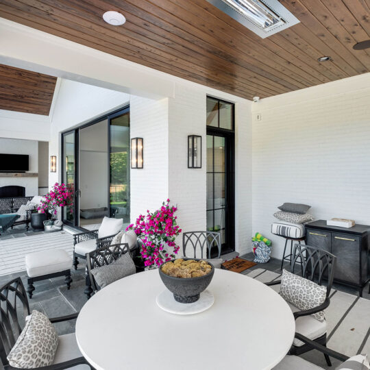 Covered patio with a round white table surrounded by black chairs. A sectional sofa with pillows is on the left, facing a wall-mounted TV. Pink flowers decorate the space. Wood paneling on the ceiling and potted plants enhance the cozy setting.