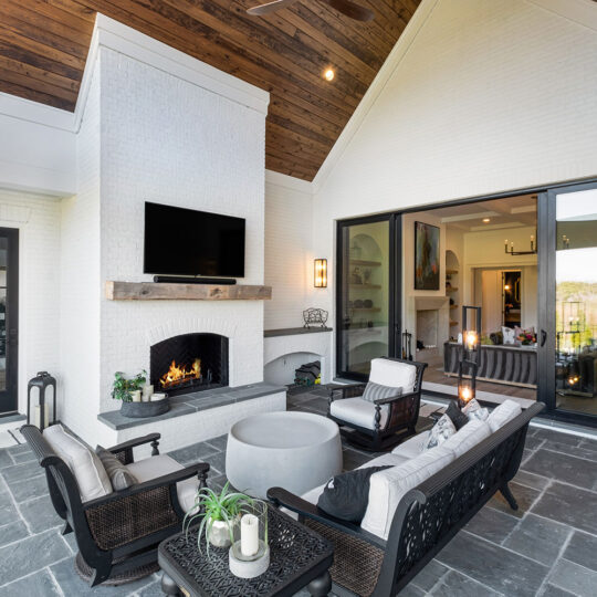 Modern outdoor living space with a cozy seating area, fireplace, and mounted TV. The patio features cushioned chairs, a stone table, and decorative lanterns. Large glass doors reveal an interior living area with artwork and natural lighting.